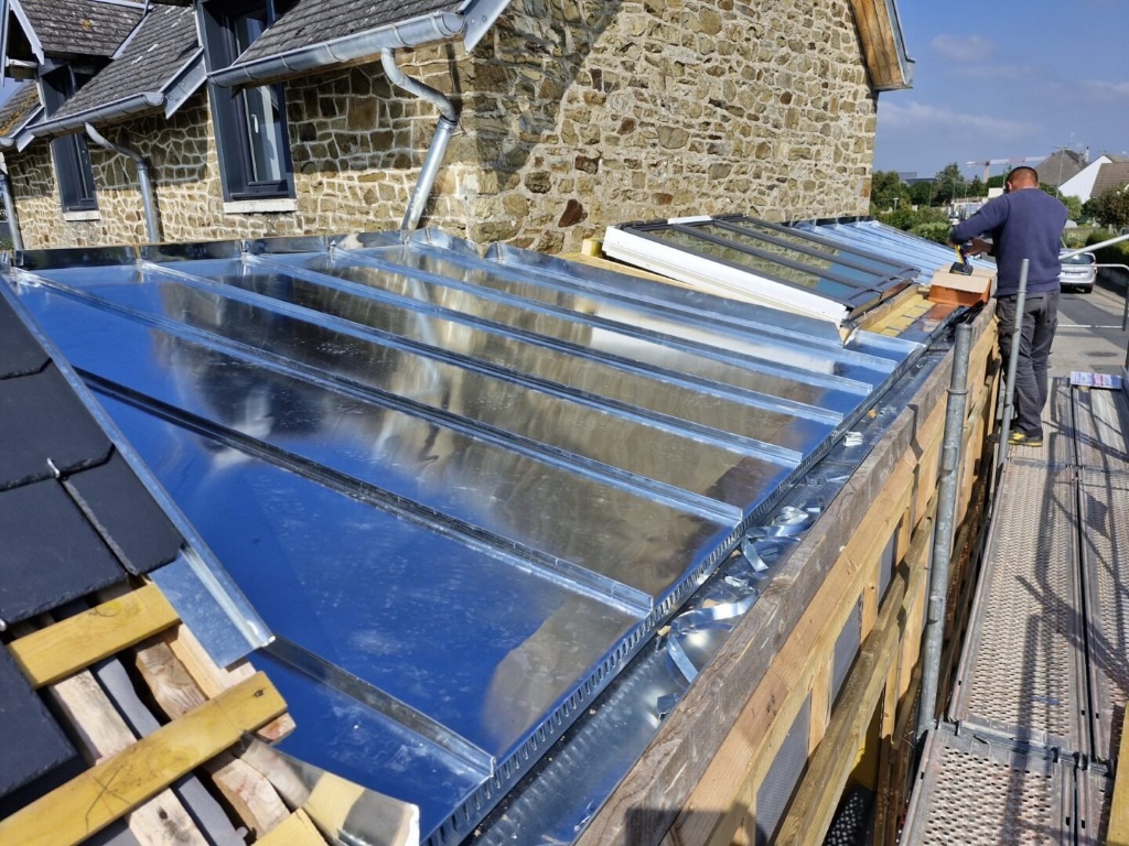 Installation d'une couverture en zinc à joint debout sur le toit d'une maison en pierre avec un ouvrier travaillant depuis un échafaudage.